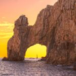 El Arco de Cabo San Lucas al atardecer, con un velero visible a través del arco rocoso y el cielo teñido de naranja y dorado
