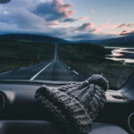Vista desde el interior de un coche con un gorro de lana en el salpicadero, frente a una carretera recta entre colinas verdes al atardecer